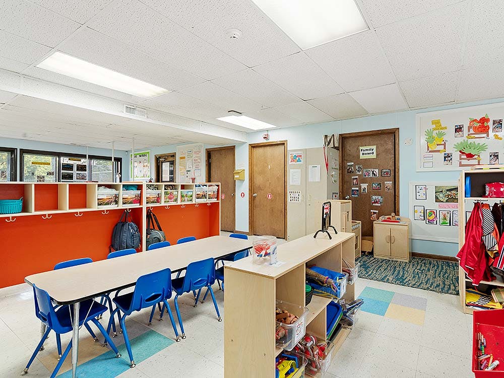Classroom with long table, blue chairs, cubbies, and play shelves