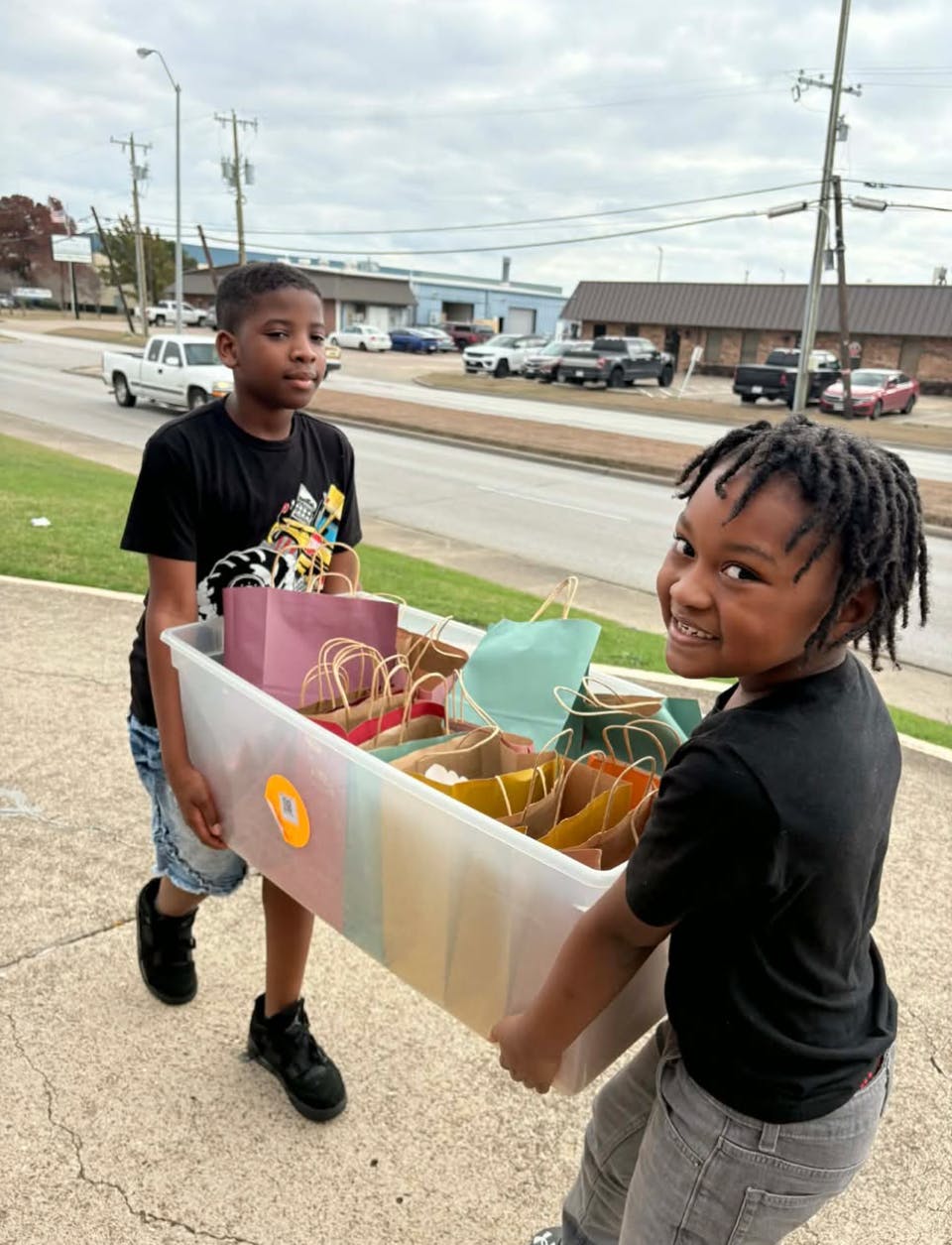 Two children carrying a large plastic bin filled with gift bags