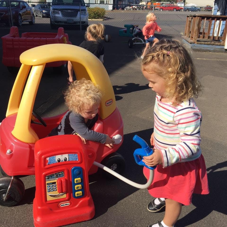 Child in toy car at toy gas pump with another child holding the nozzle