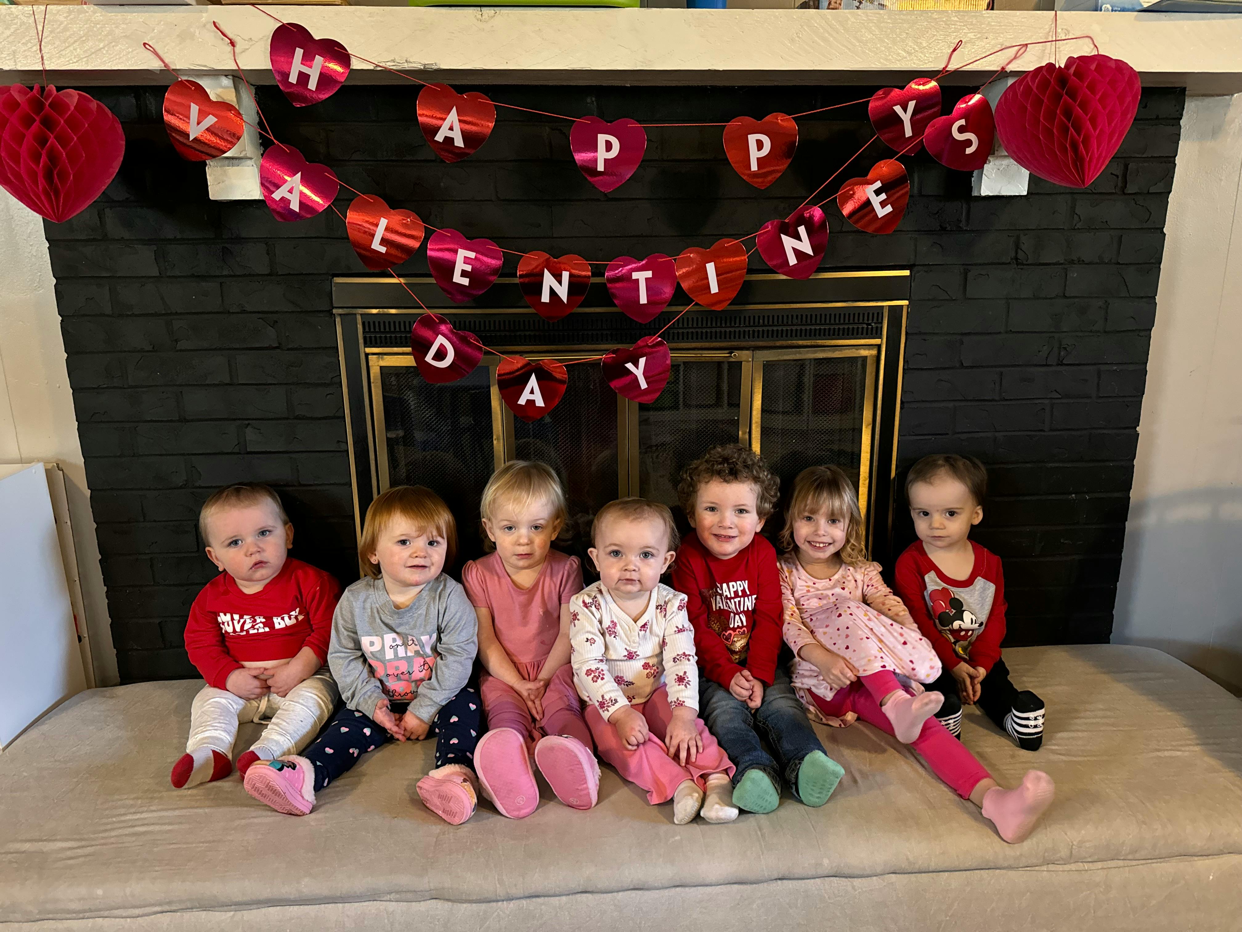 Seven children sitting in front of a fireplace with a "Happy Valentine's Day" banner