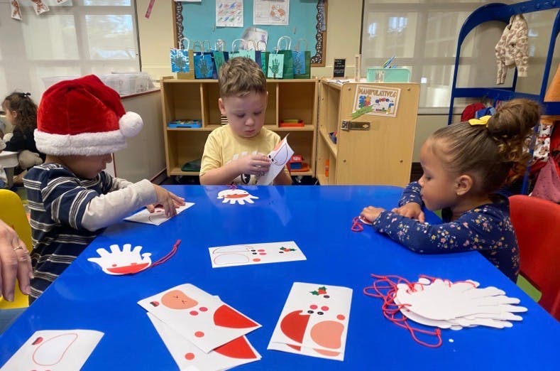 Three children doing crafts with paper cutouts at a table in a classroom
