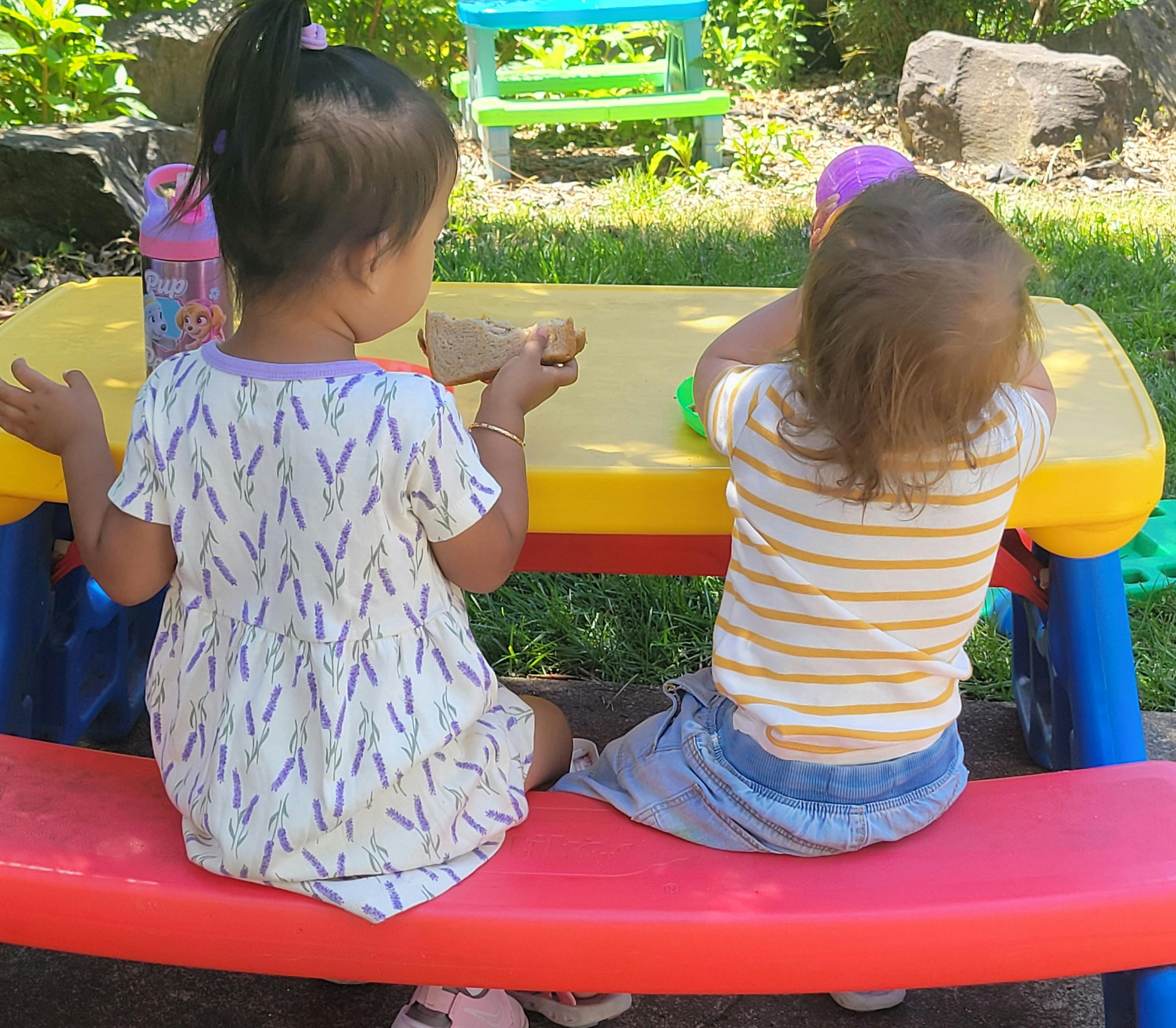 Two young children sitting at a small picnic table outdoors, one eating a sandwich