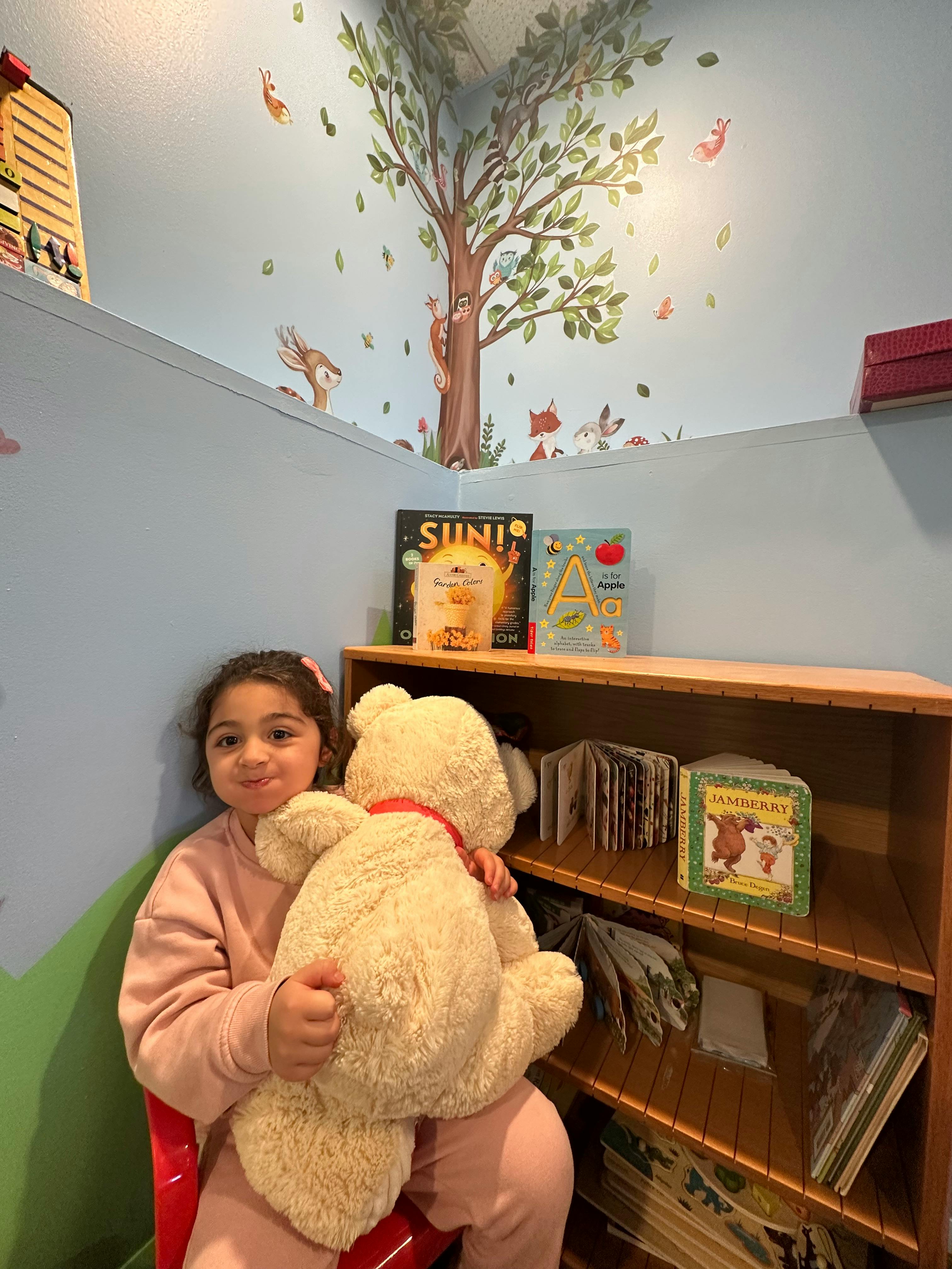 Child holding a large teddy bear in a room with a tree mural
