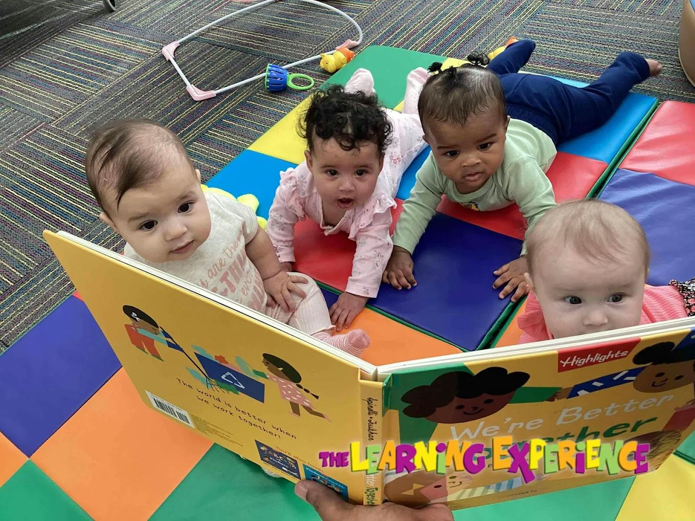 Four babies on a colorful mat looking at a book held by an adult