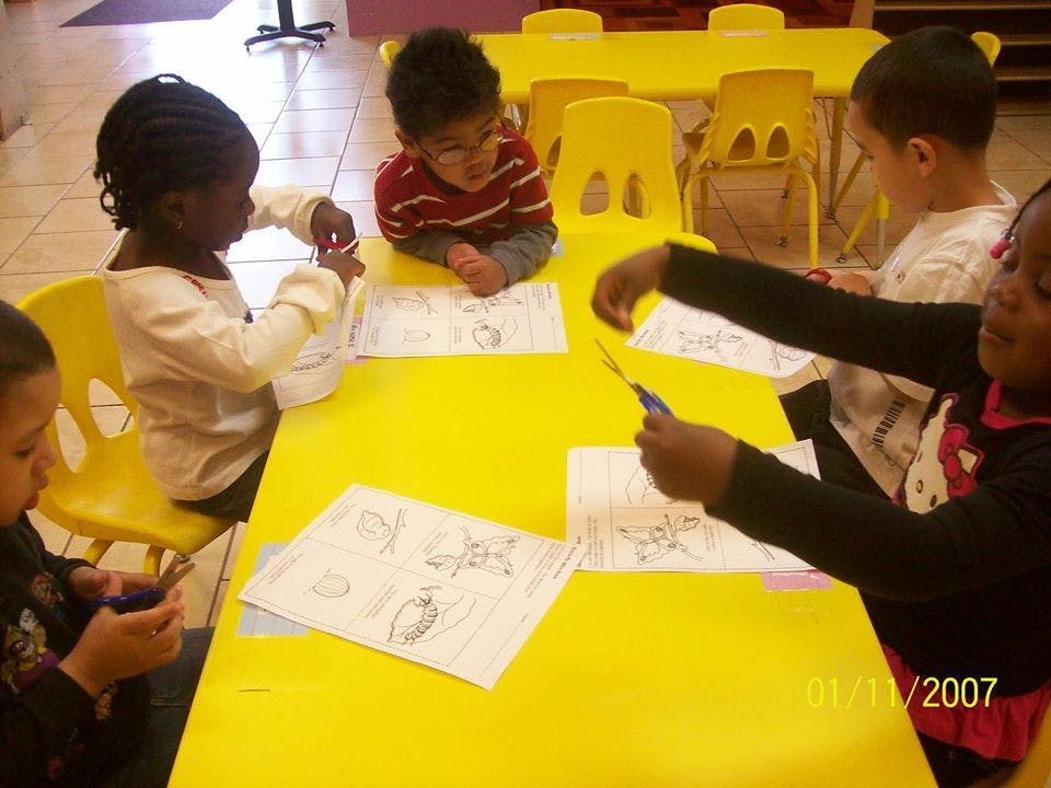 Four children cutting paper at a yellow table in a classroom