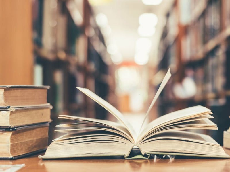 An open book on a table with more books stacked next to it, library shelves in background