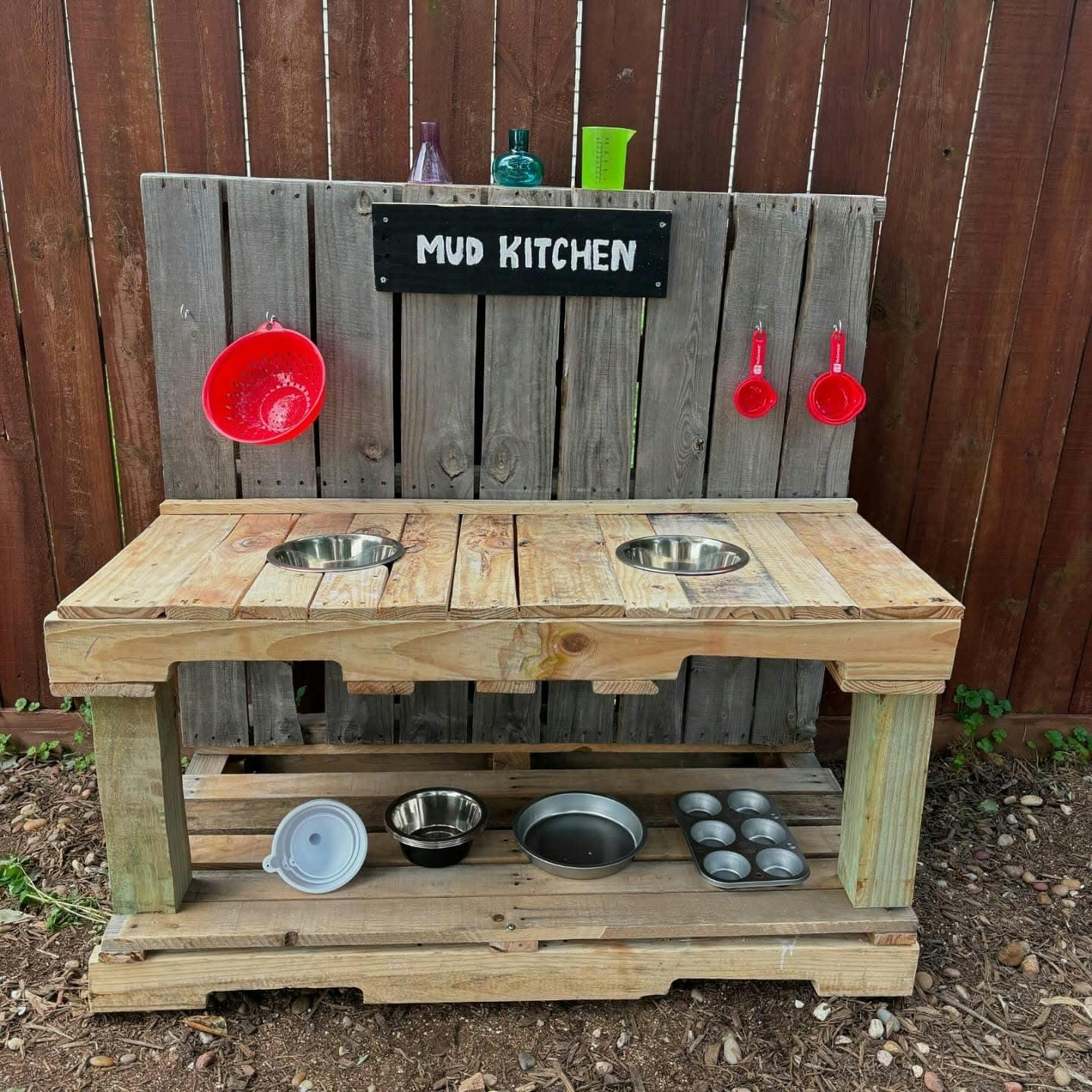 Wooden mud kitchen with a "Mud Kitchen" sign, bowls, and utensils