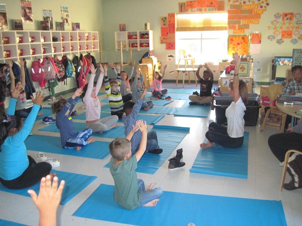 Children and adults sitting on mats with arms raised in a classroom