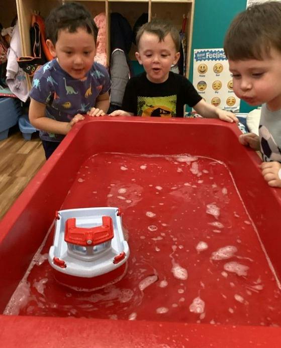 Three children playing with a toy boat in a red bin with soapy water