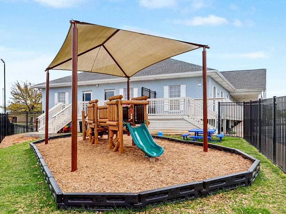 Playground with wooden play structure, green slide, and sunshade on wood chips