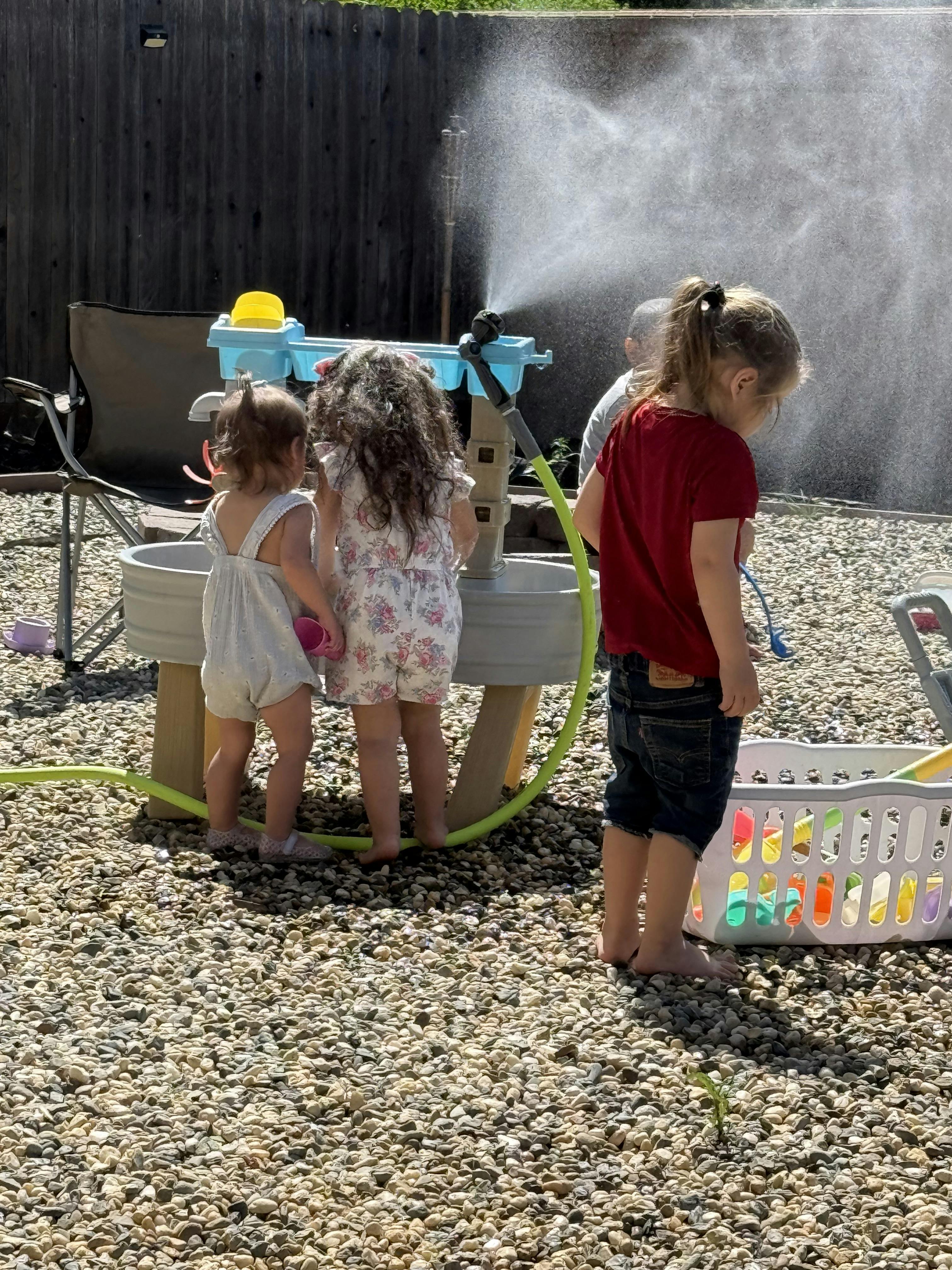 Three children playing outdoors with a water table and sprinkler