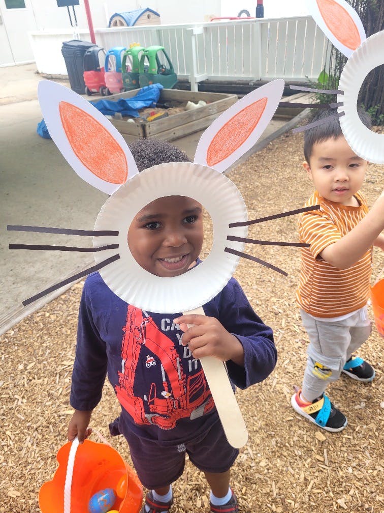 Child wearing a paper plate bunny mask, holding a craft stick and a basket