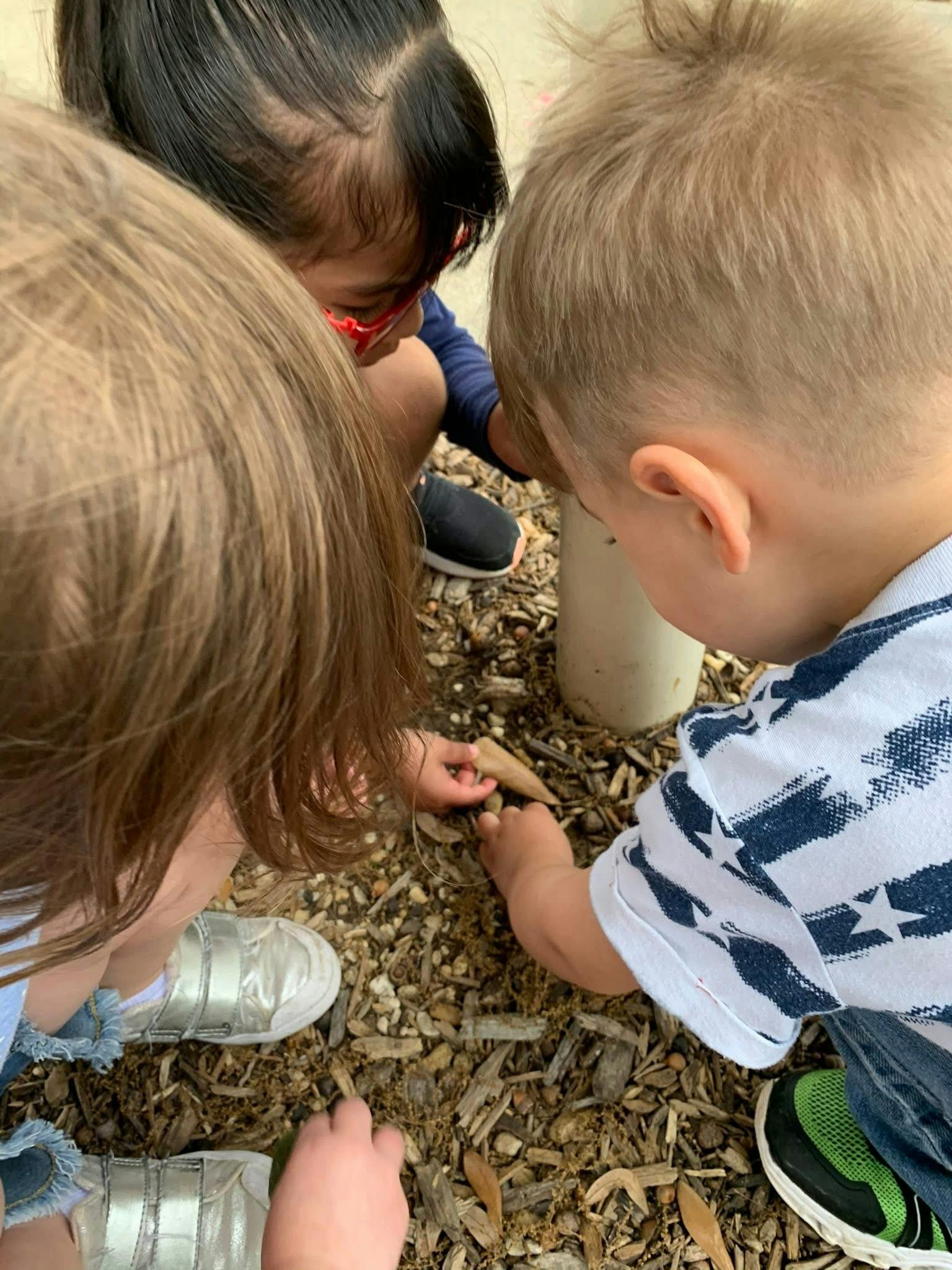 Three children playing outdoors, looking down at the ground