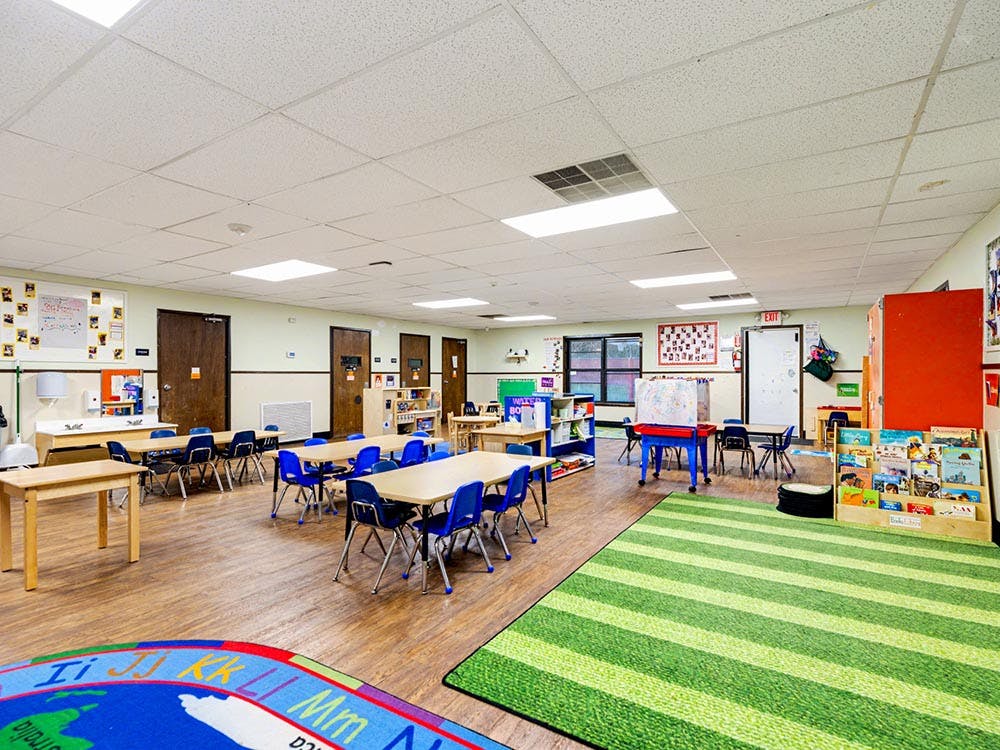 Empty classroom with small tables and chairs, and a striped green rug