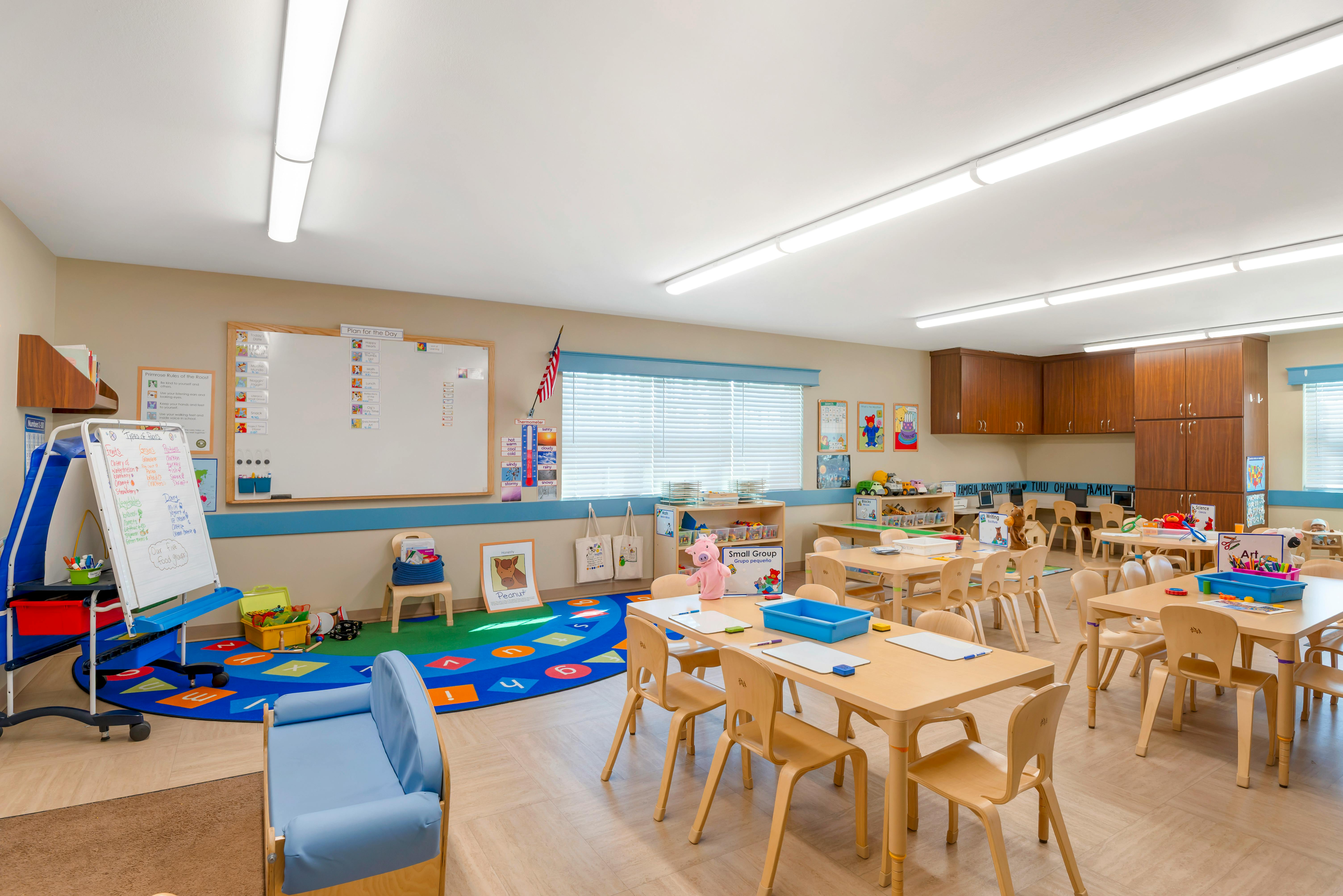 Empty classroom with small tables and chairs, a whiteboard, and a rug with letters