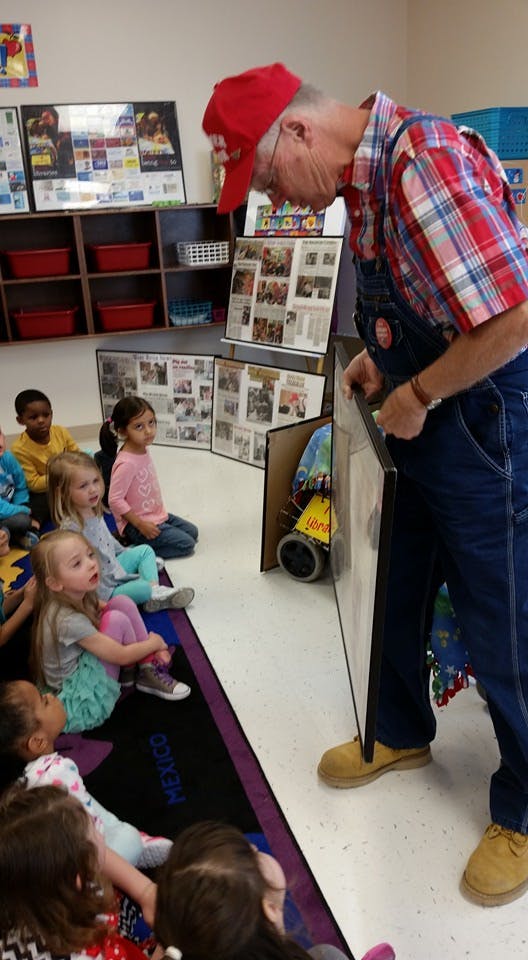 Person in red hat and overalls showing something to children sitting on floor