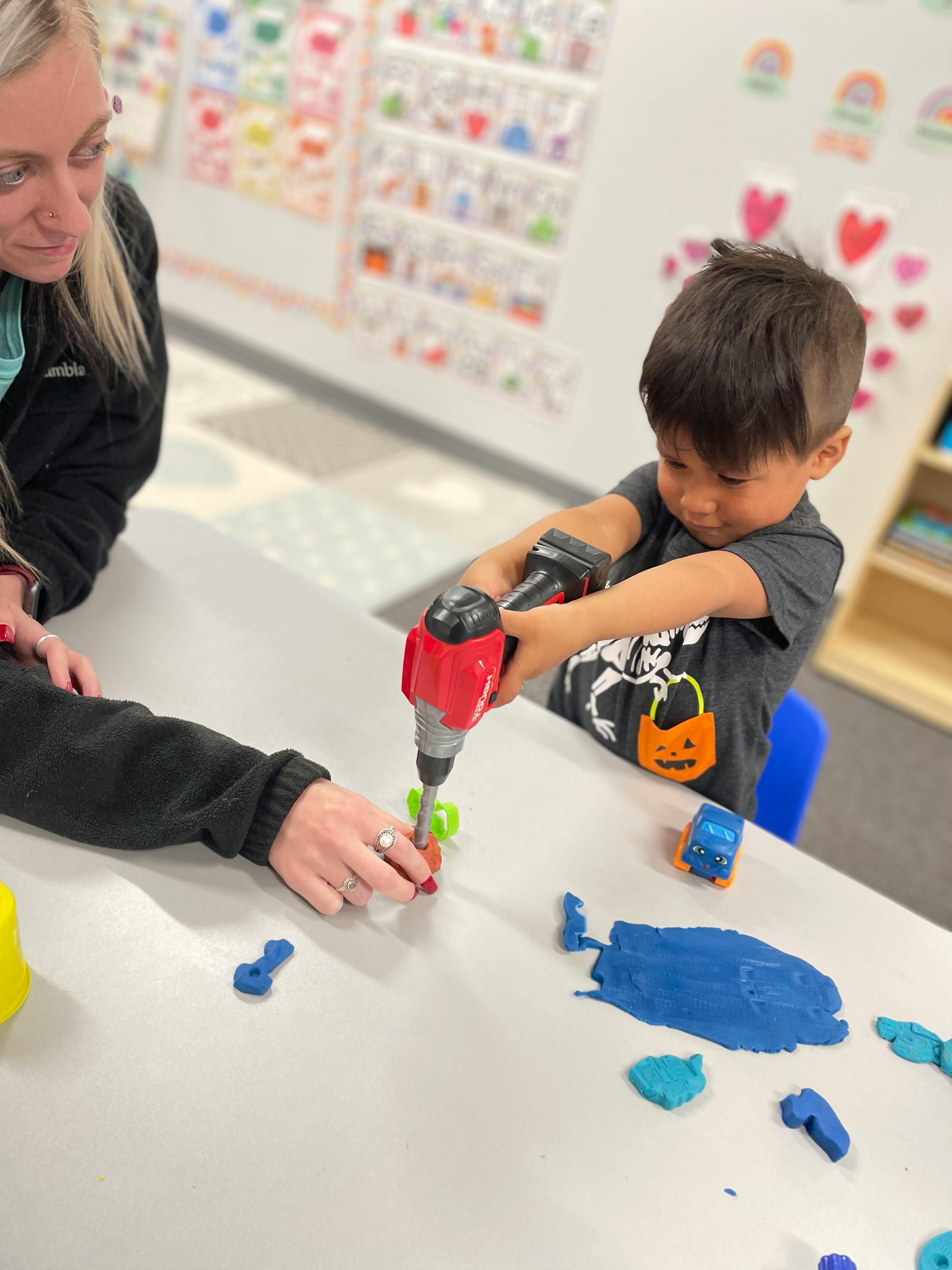 Adult and child playing with toy drill and playdough in a classroom