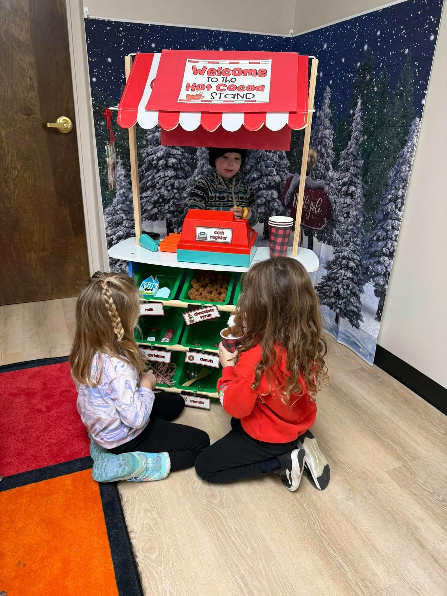 Child at a play hot cocoa stand with two children sitting in front