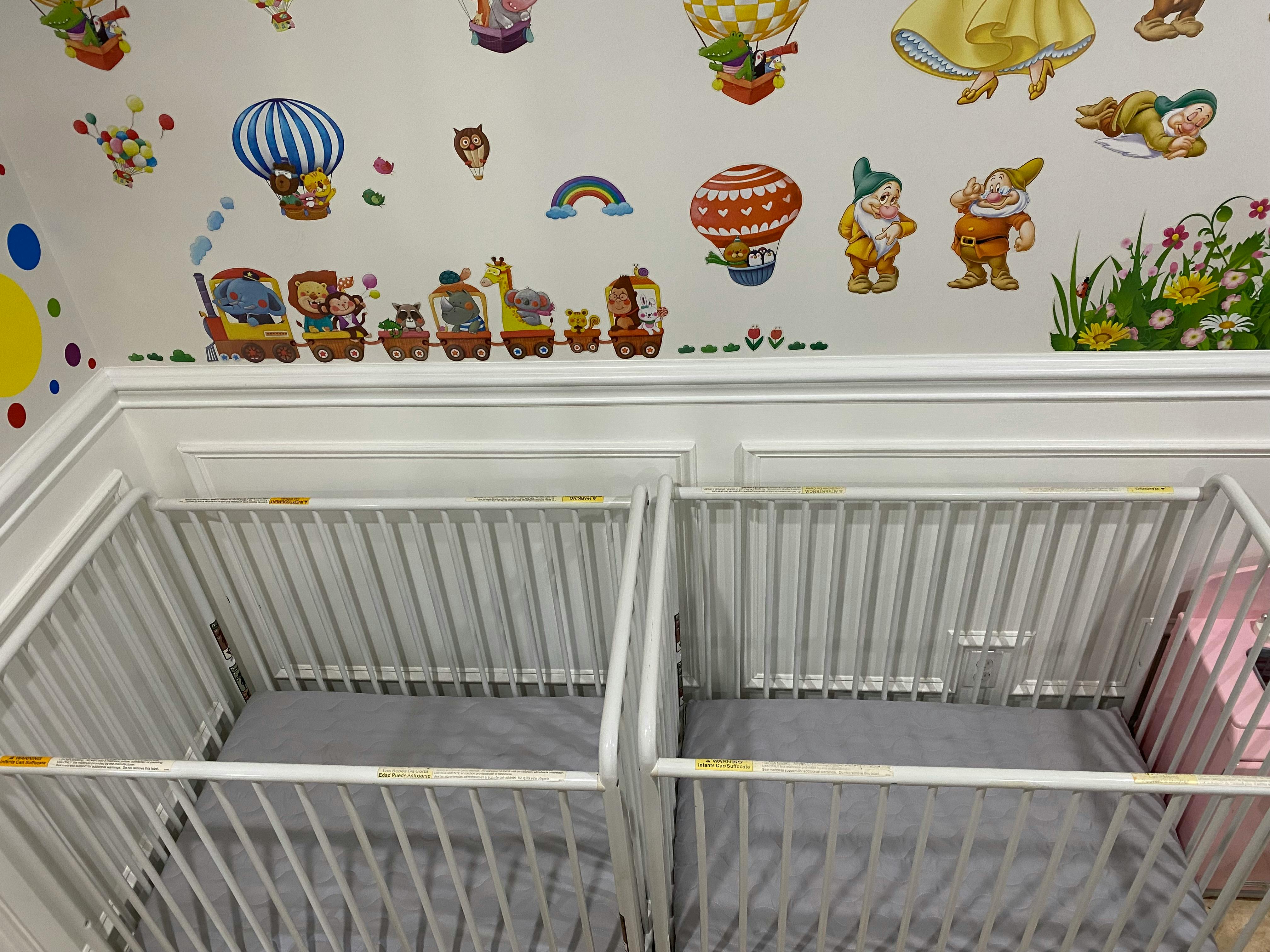 Two empty white cribs with gray sheets in a room with wall decals
