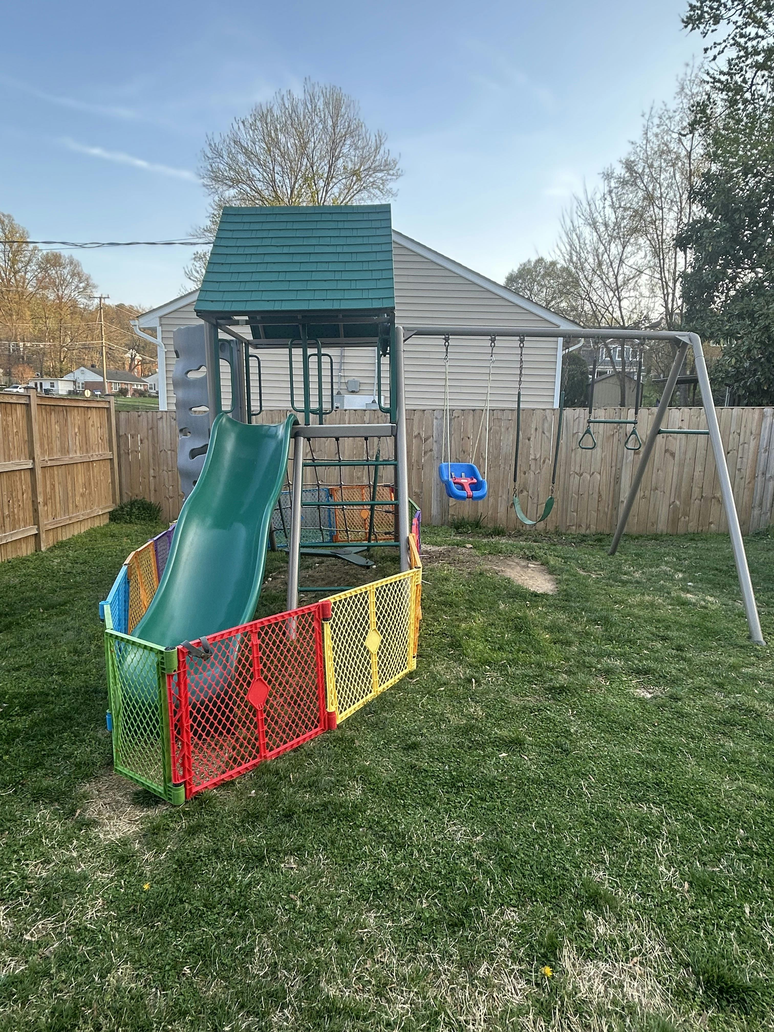 Playground set with green slide, swings, and colorful safety fencing in a grassy backyard