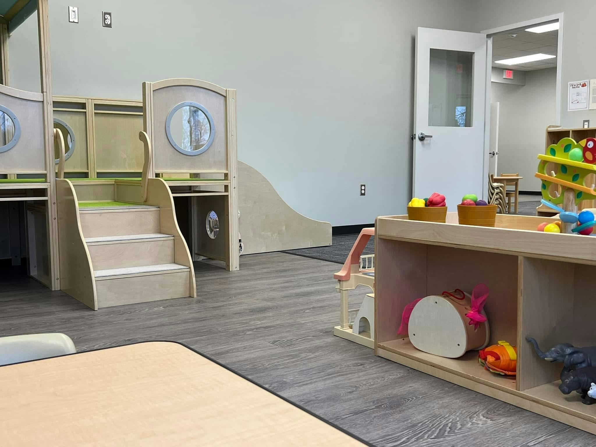 Childcare classroom with wooden play structures, shelves of toys, and open doorway