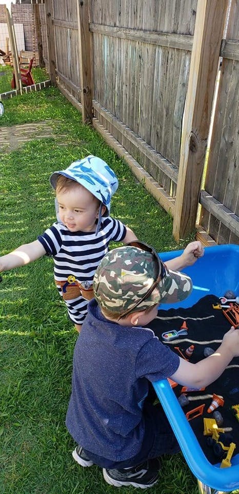 Two children playing outdoors, one standing and one kneeling by a blue bin