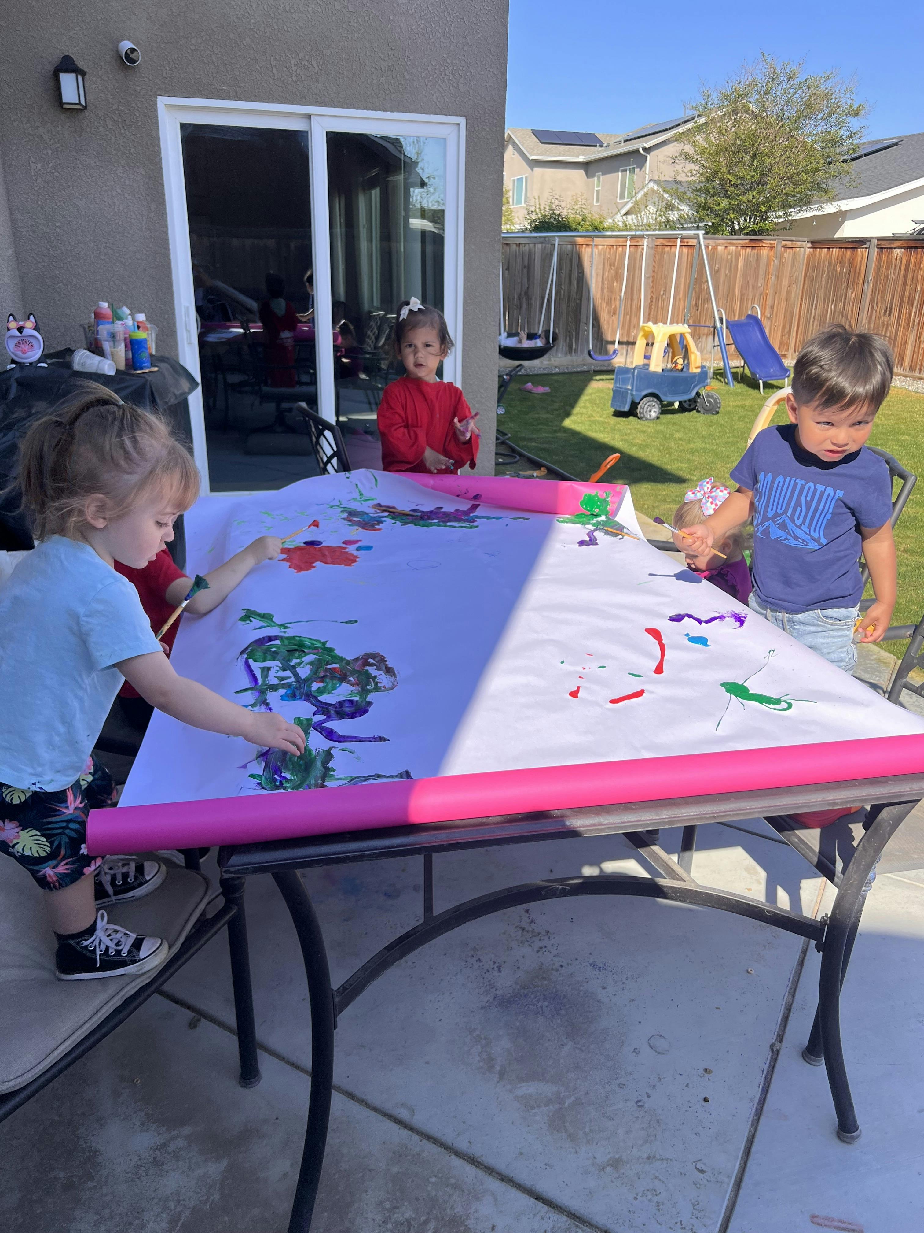 Three children painting on a large white sheet on a table outdoors
