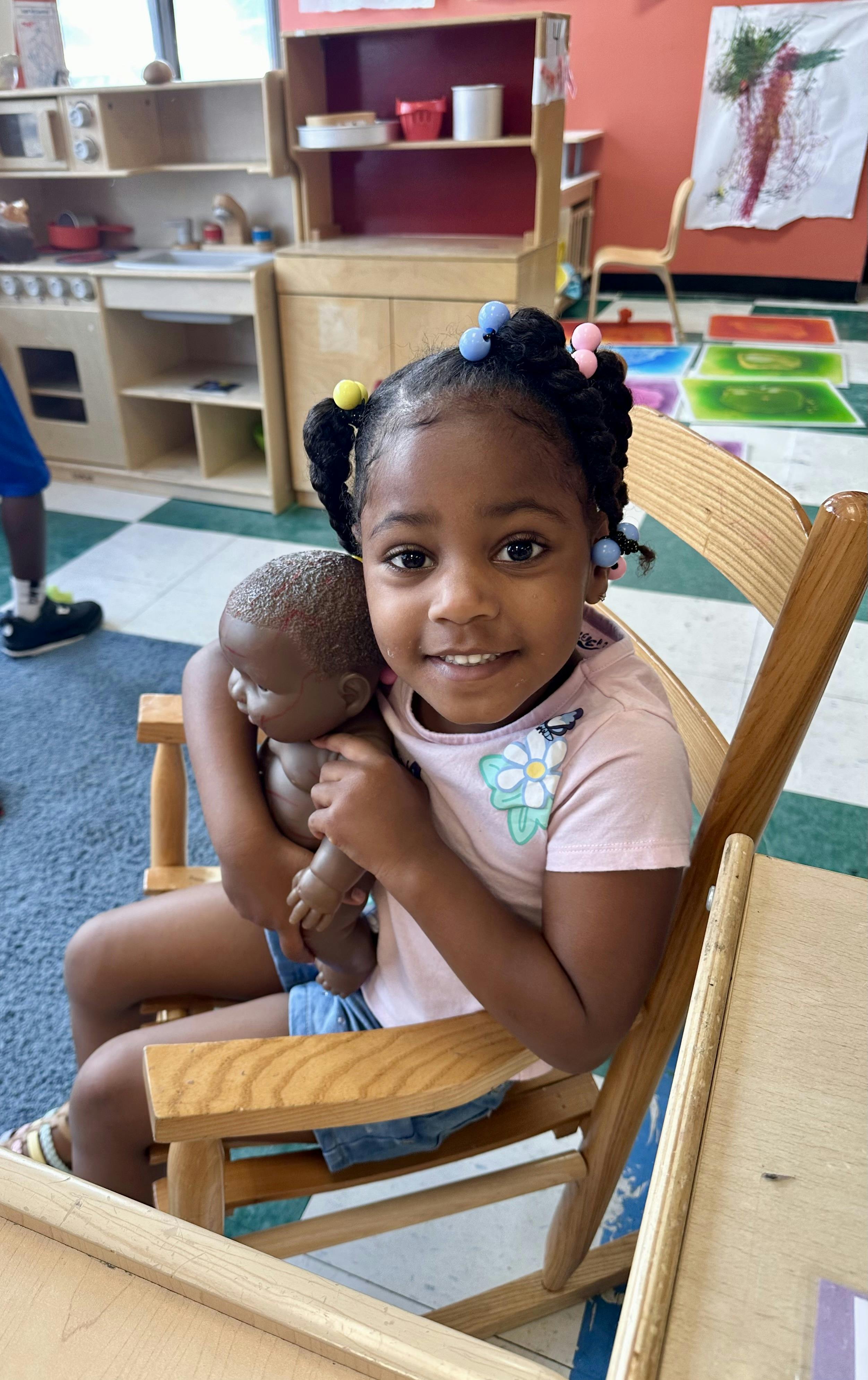 Child sitting in a wooden chair holding a doll in a classroom