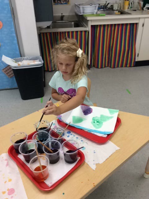 Child painting with multiple cups of paint on a table in a classroom