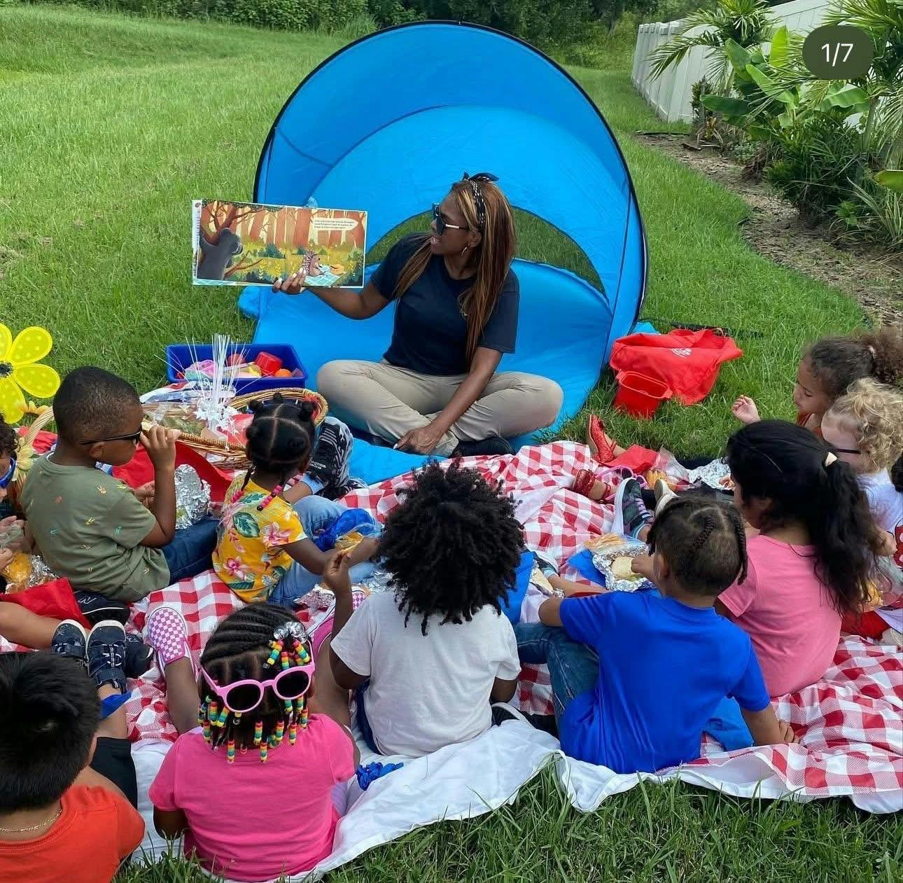 Person reading a book to a group of children seated on a picnic blanket outdoors
