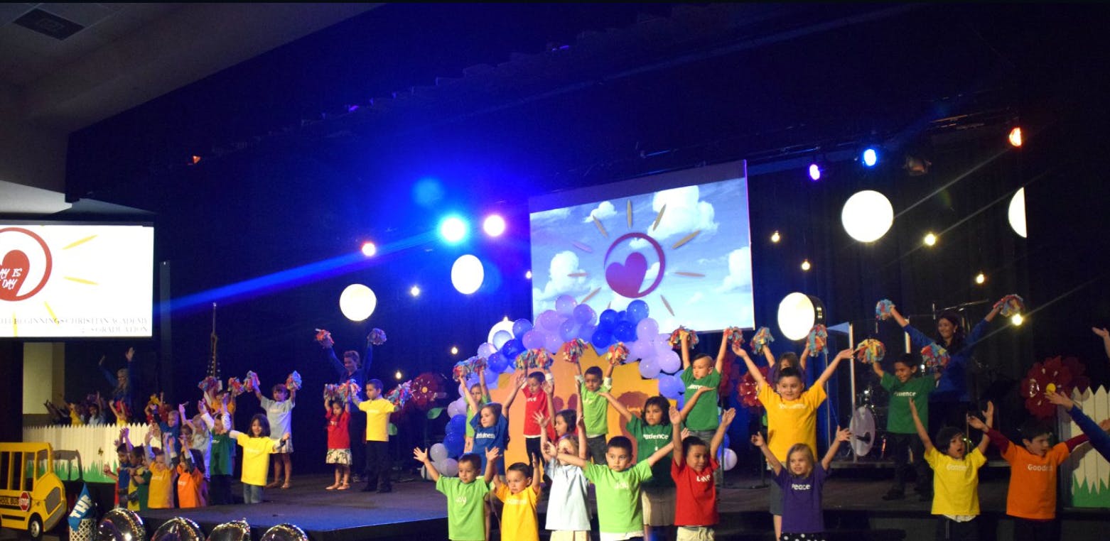 Children on a stage with arms raised, a screen displaying a heart in the background