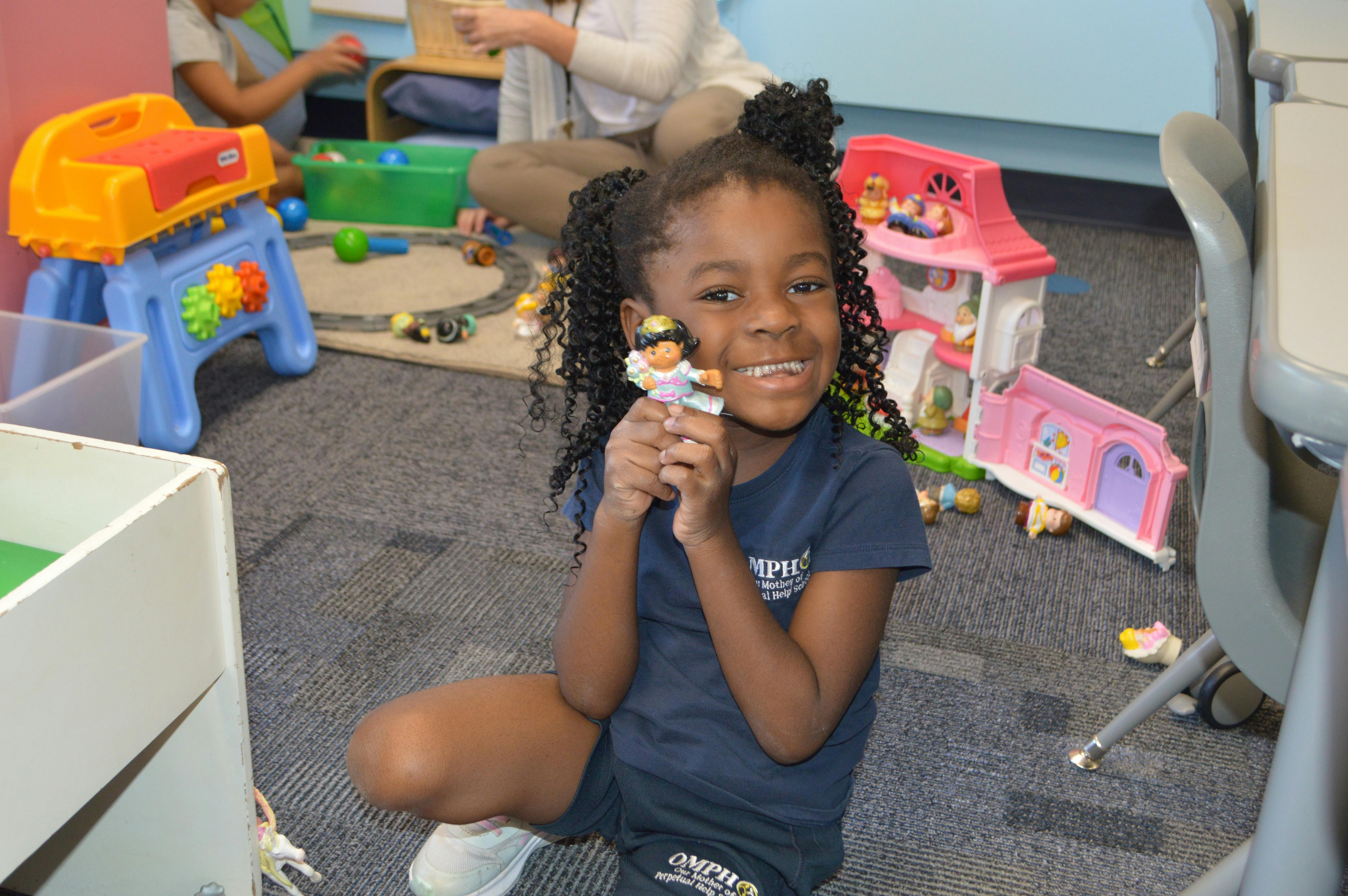 Child holding a small doll while sitting on the floor in a room with toys