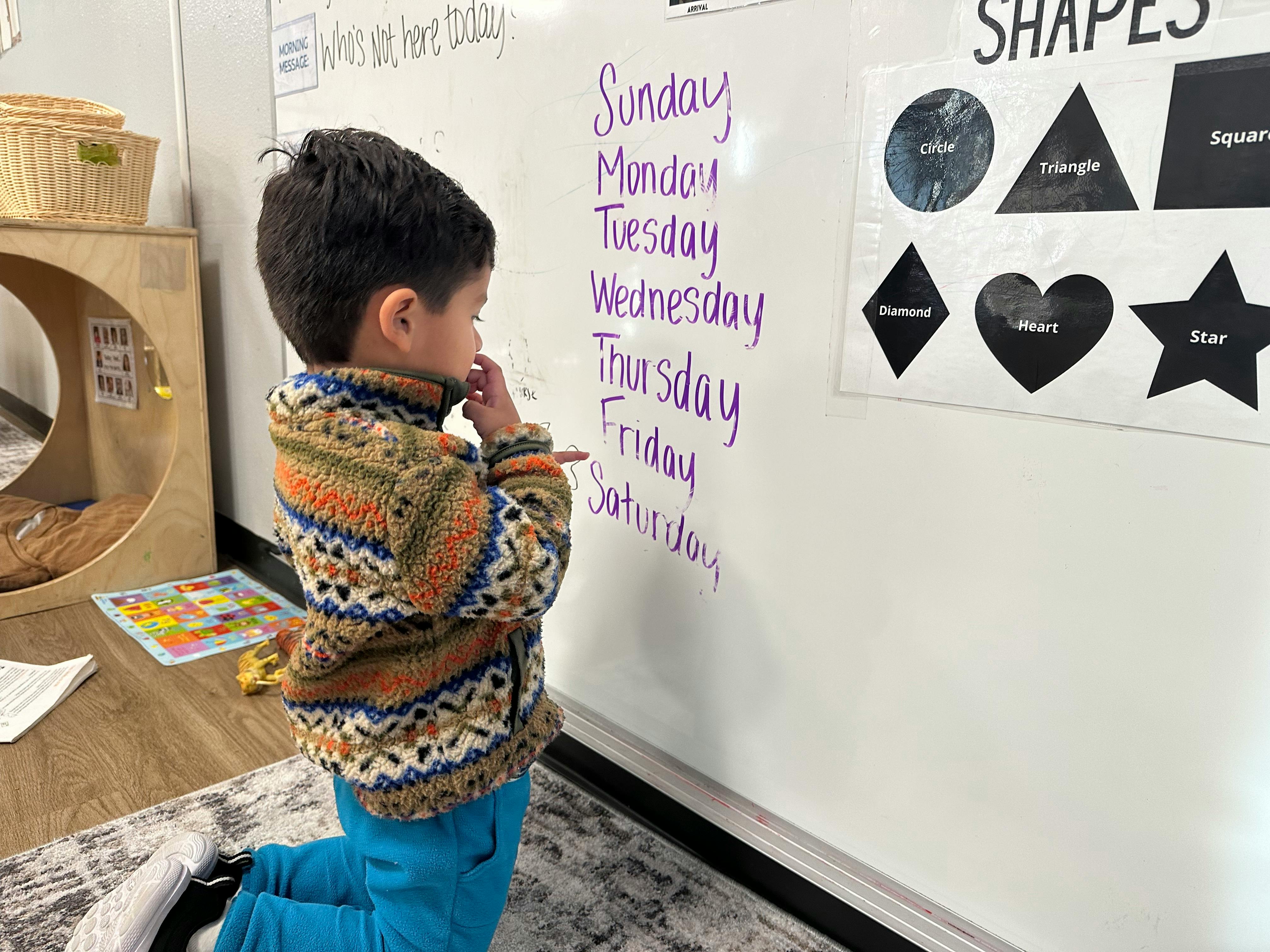 Child kneeling and pointing at a whiteboard with days of the week written on it