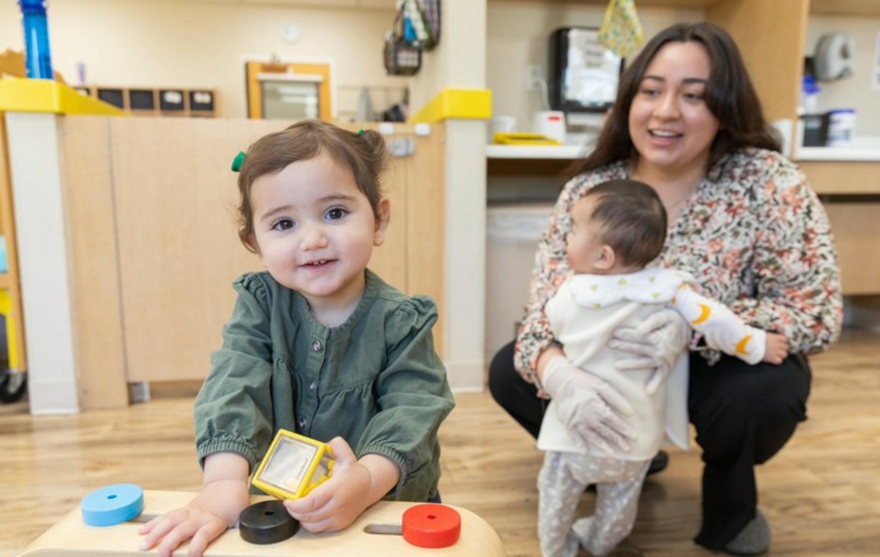 Smiling child playing with blocks as an adult holds an infant in the background