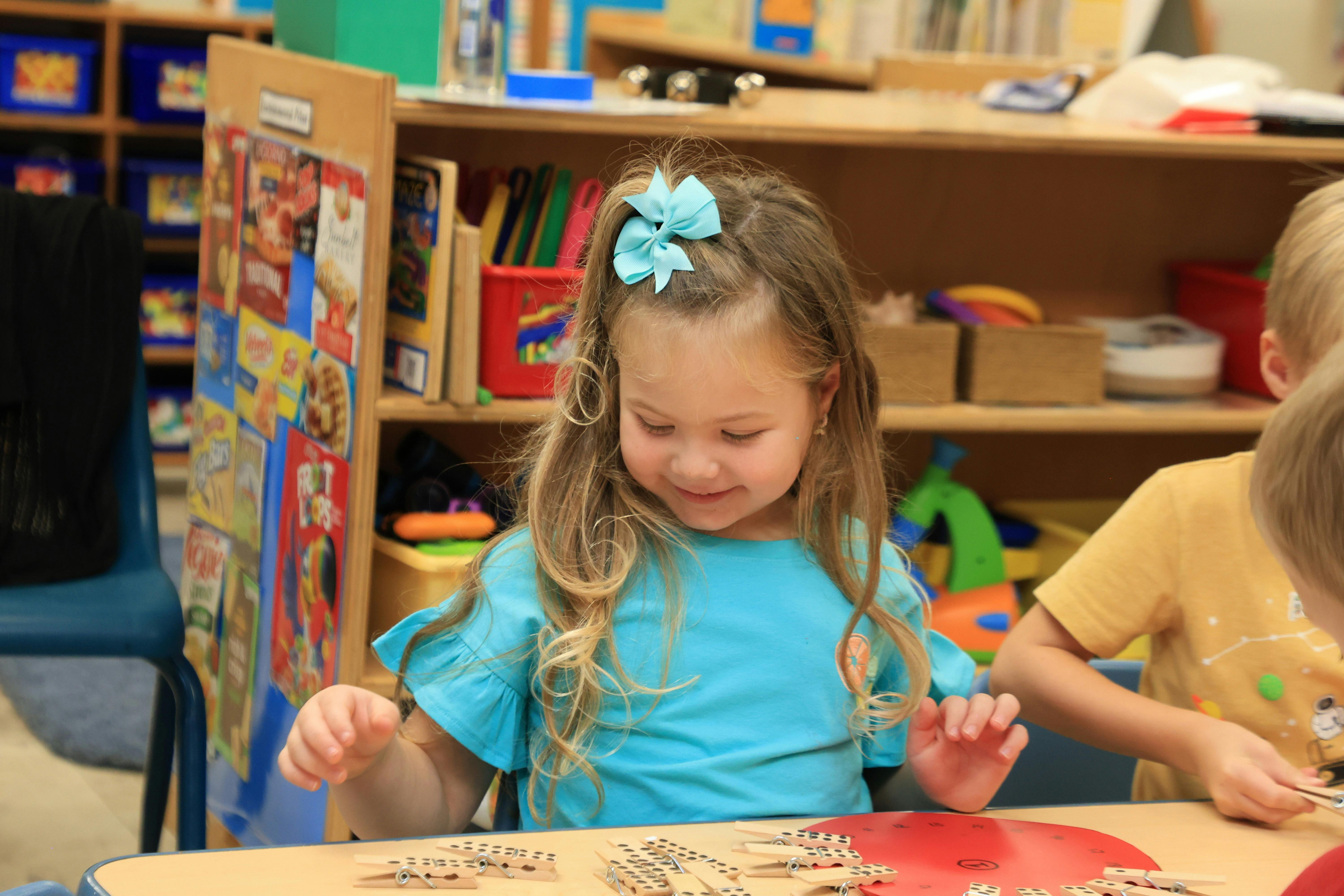 Child with a blue bow in hair looking down at a table in a classroom