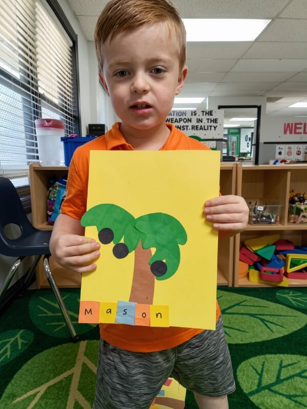 Child holding a yellow paper with a paper cutout of a tree labeled Mason