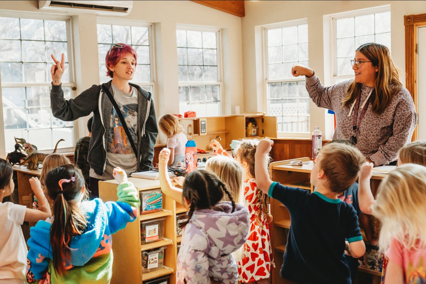 Two people teaching a group of children raising their fists in a classroom
