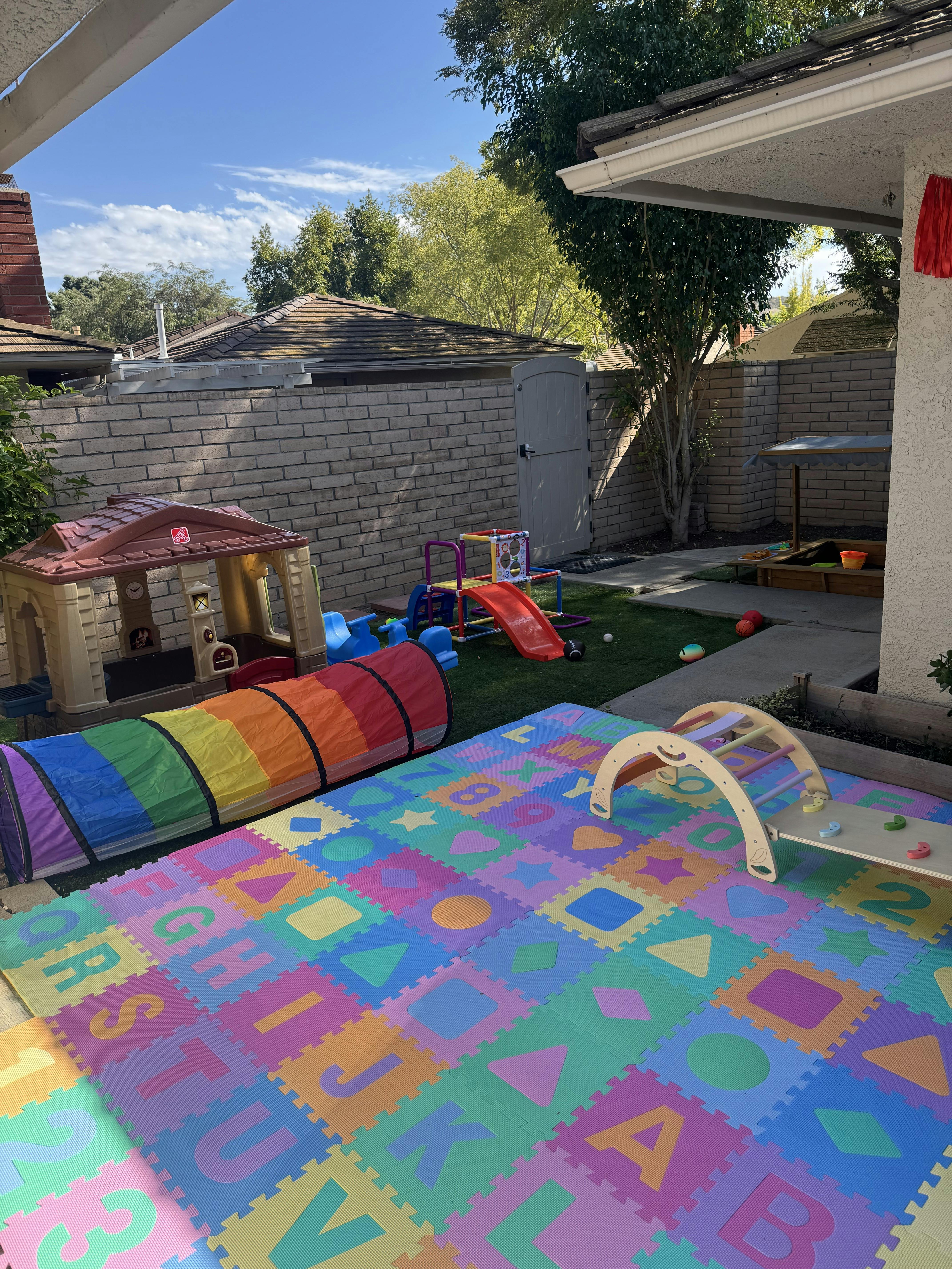 Outdoor play area with a rainbow tunnel, playhouse, and alphabet mat