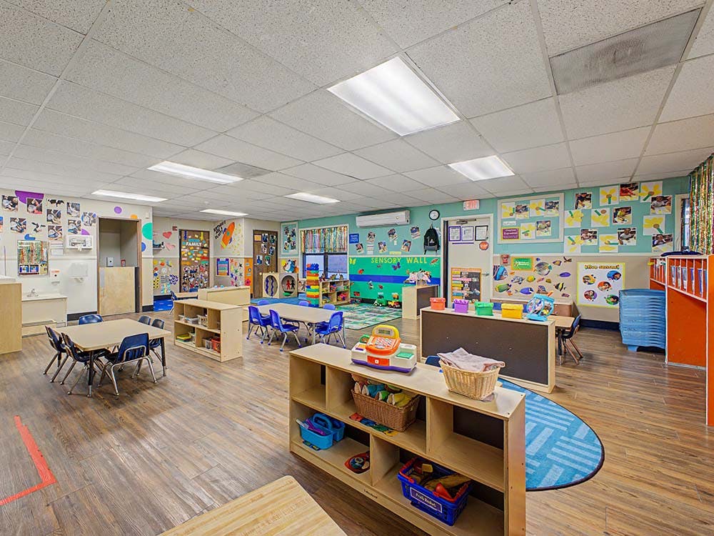 An empty classroom with small tables and chairs, shelves of toys, and decorated walls