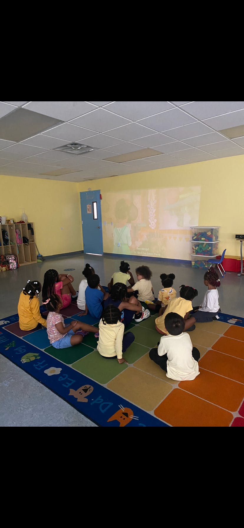 Children sitting on a colorful rug in a classroom, looking at a projected image