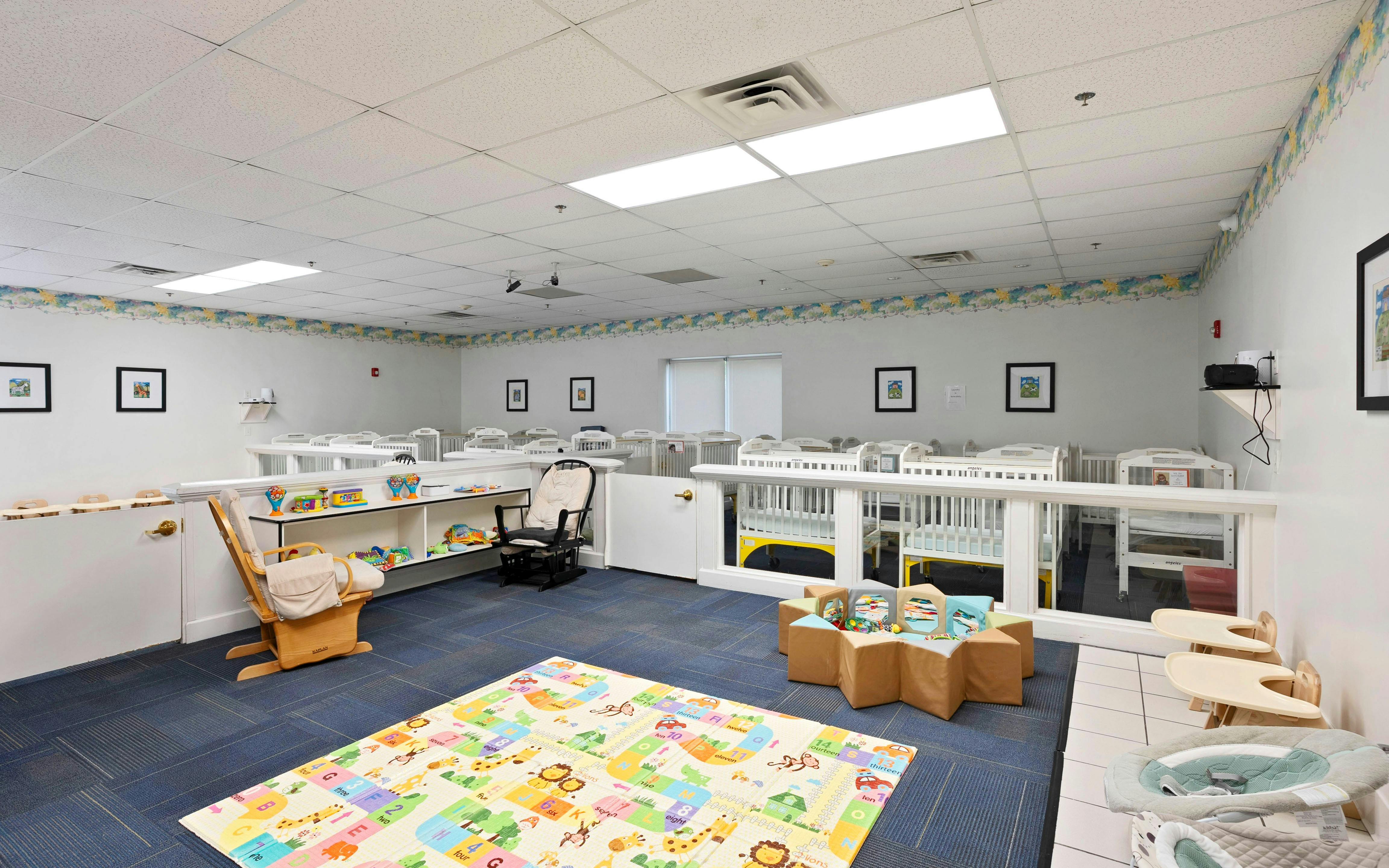A daycare room with many white cribs, a playmat on the floor, and a rocking chair