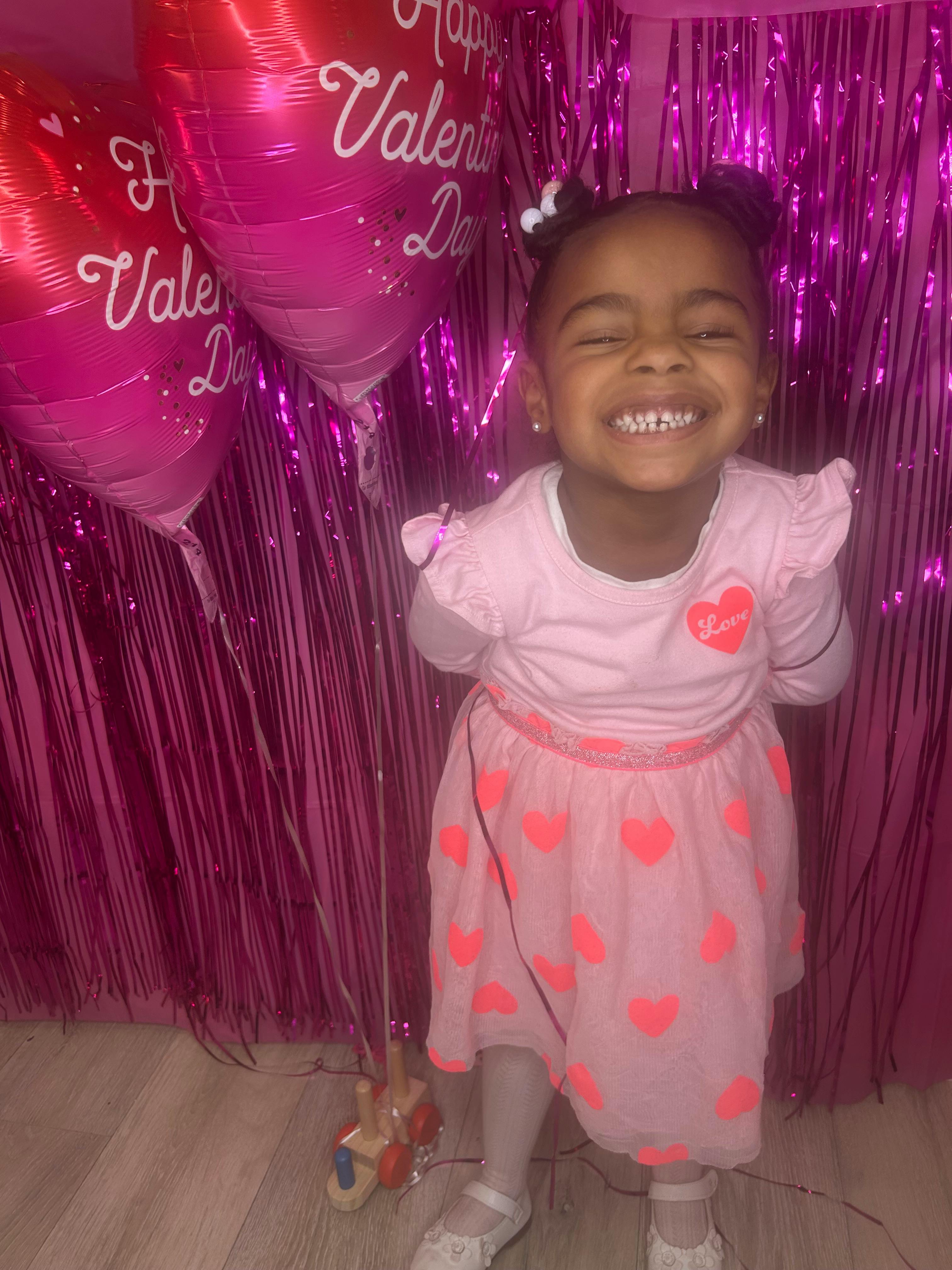 Child in pink dress with hearts, smiling in front of pink balloons and purple streamers