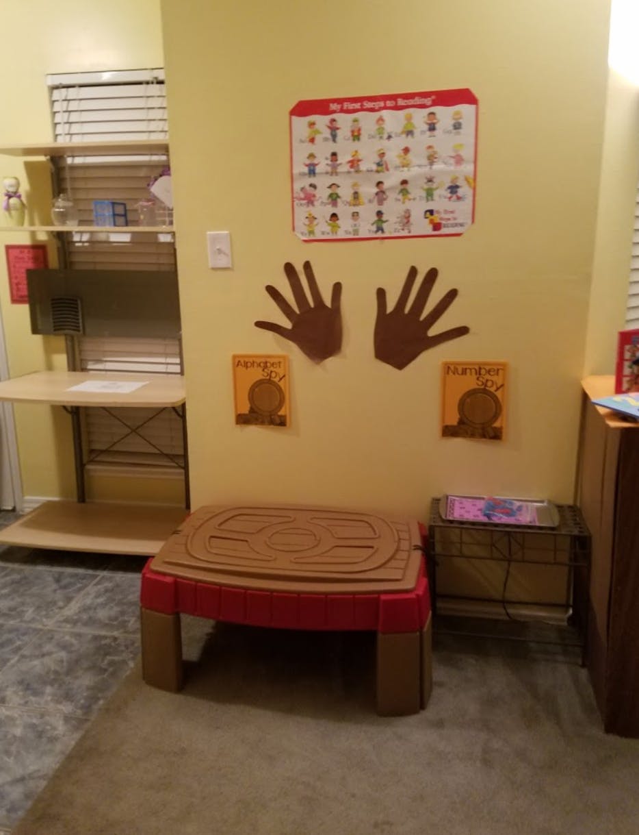 Classroom wall with a chart, paper hand cutouts, and a child's play table