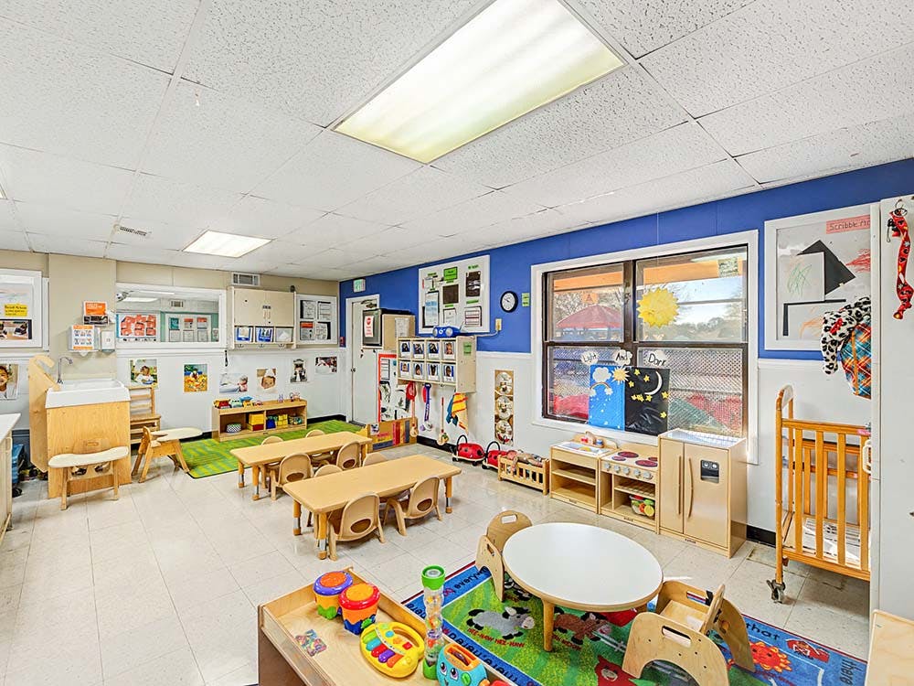 Empty classroom with small tables and chairs, play kitchen, and colorful rugs