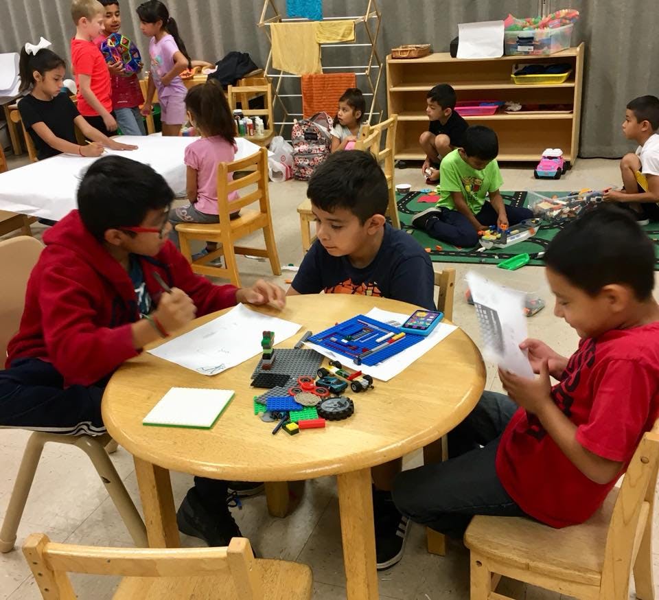 Children in a classroom, some sitting at tables, others on the floor