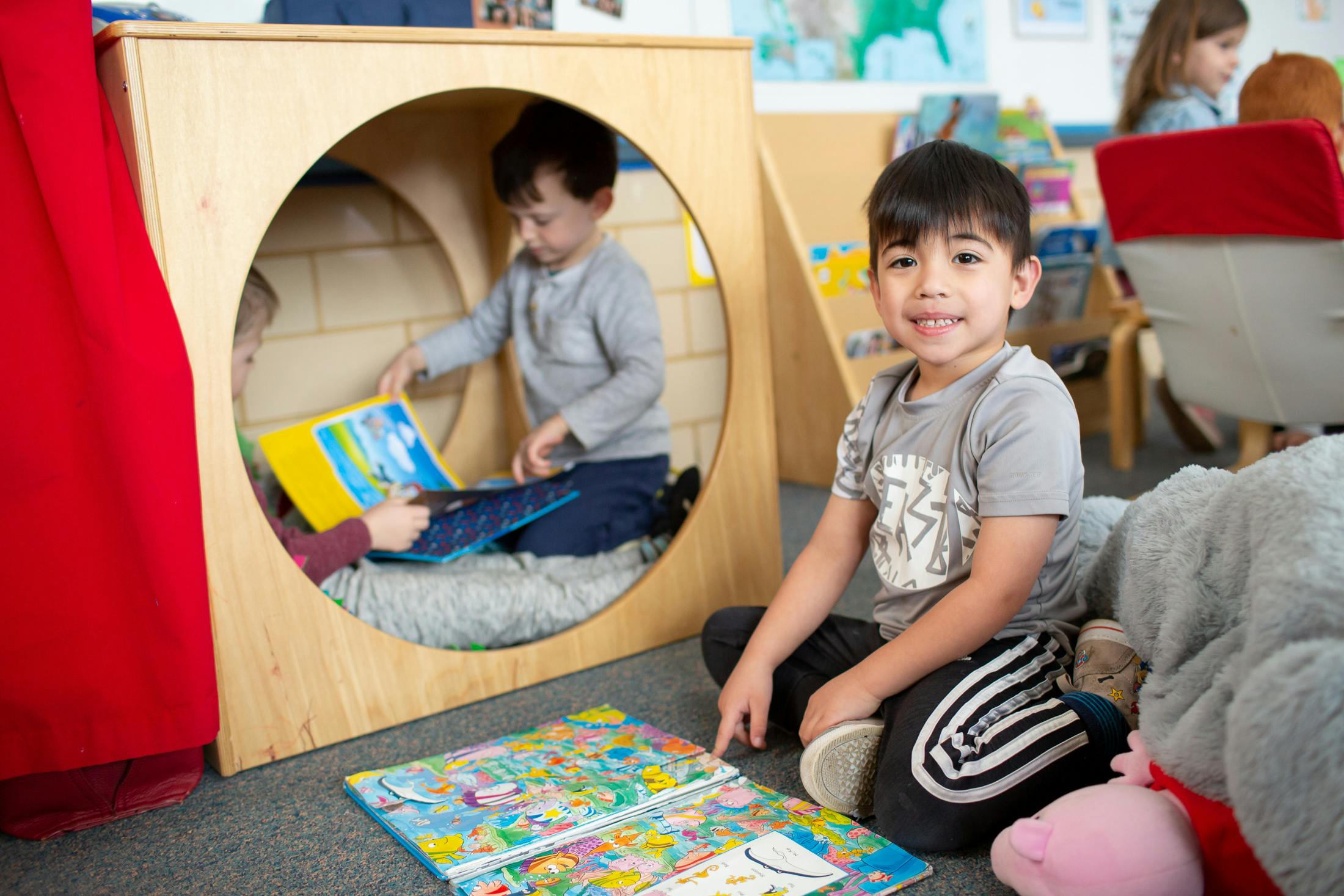 Child sitting on floor with open book, another child reading in a wooden cubby
