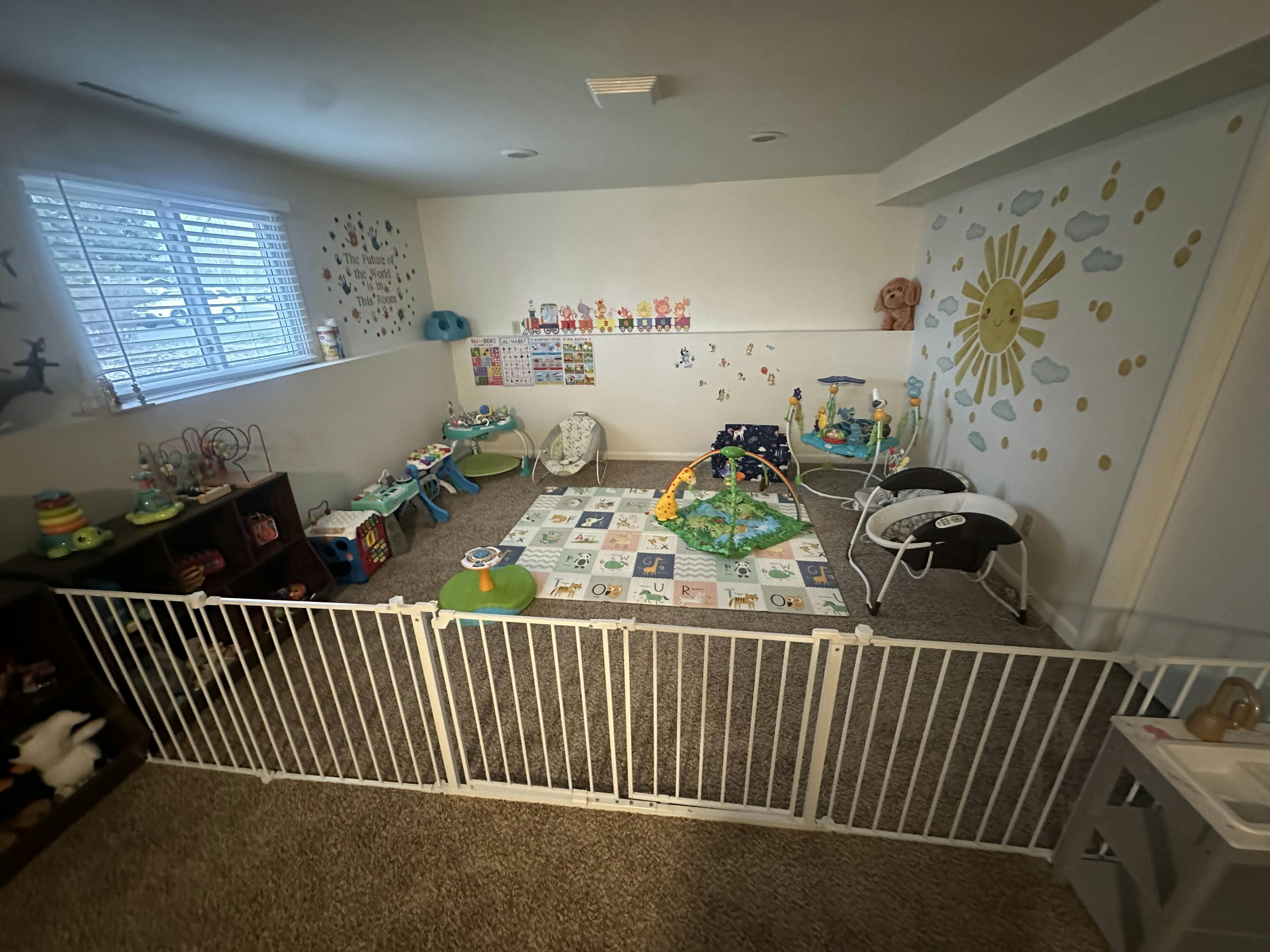 A room with baby toys, playmats, a swing, and a playpen with a mural on the wall
