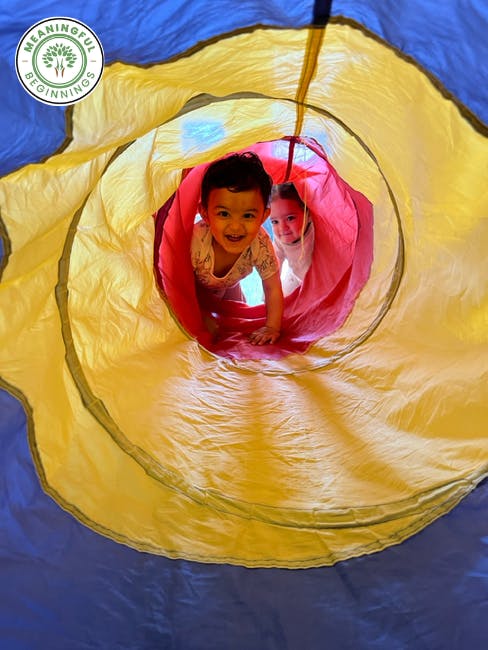 Smiling child crawling through a red play tunnel, another child behind