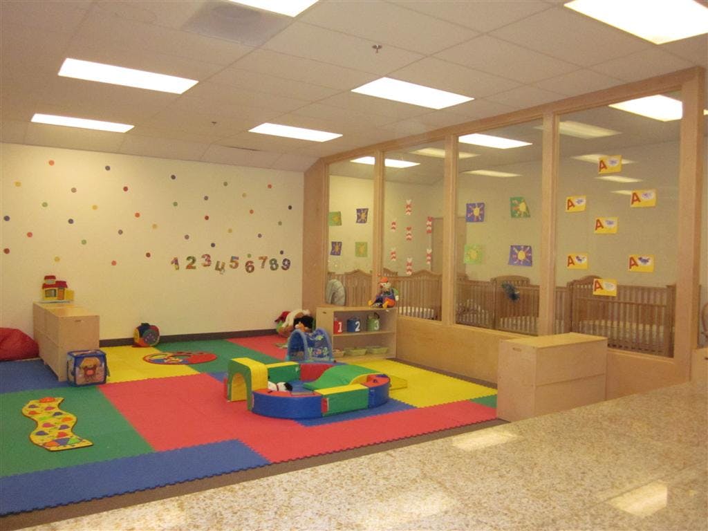 Child playing in a colorful play area with cribs visible through a glass partition
