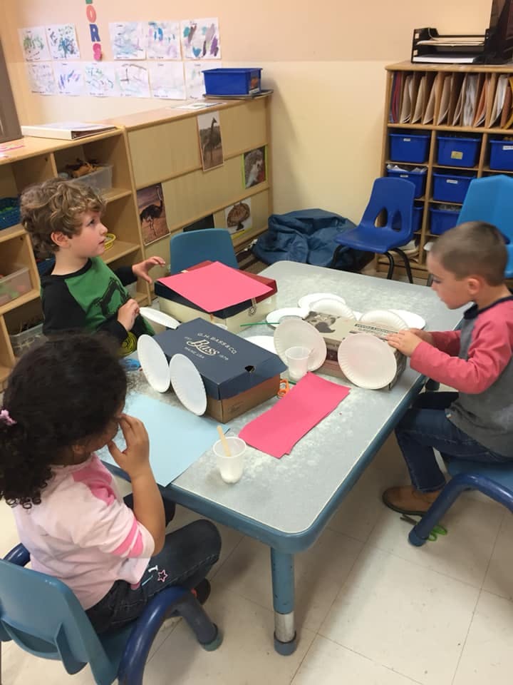 Three children at a table making crafts with paper plates and boxes